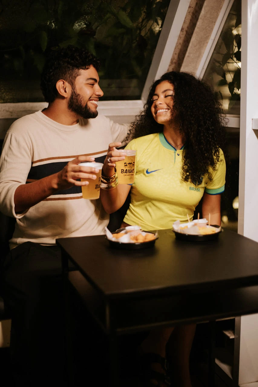 Casal sorrindo brindando copos do Baiúca com petiscos na mesa